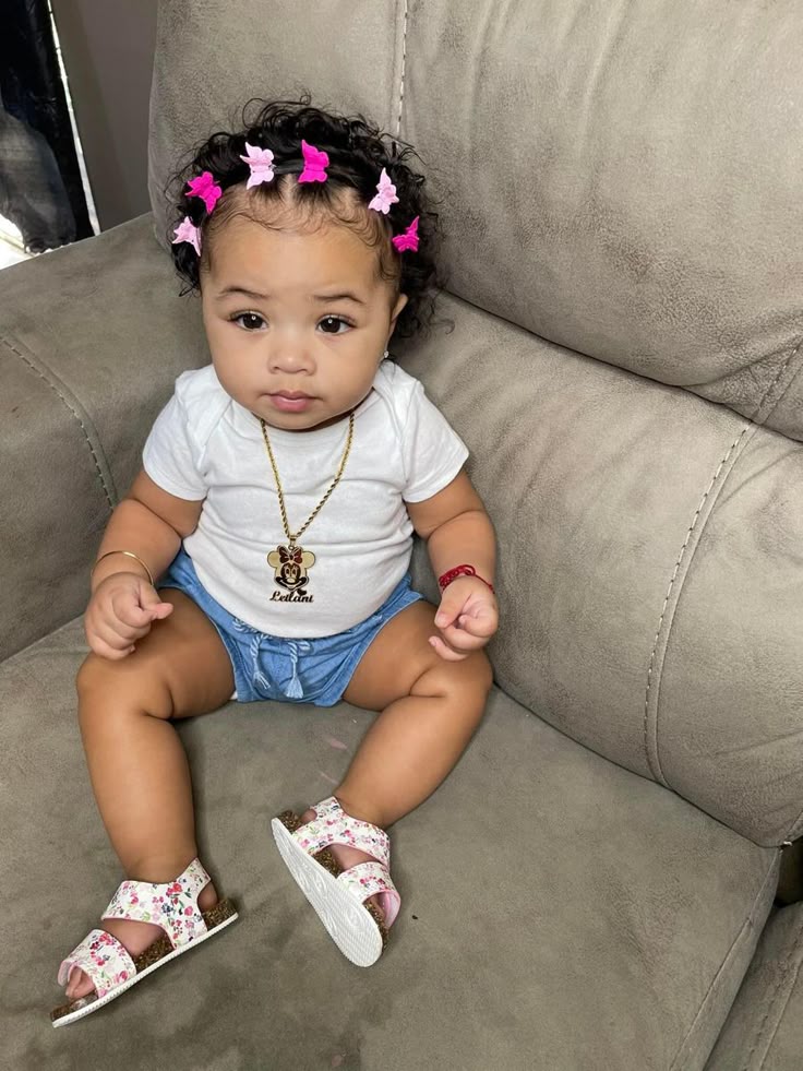 "A toddler with curly hair styled into small twists, decorated with pink bow hair clips, sitting on a couch wearing sandals."
