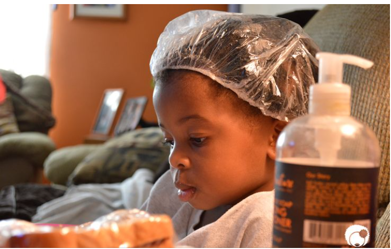 Young child wearing a plastic cap during a deep conditioning hair treatment, conditioner bottle nearby.