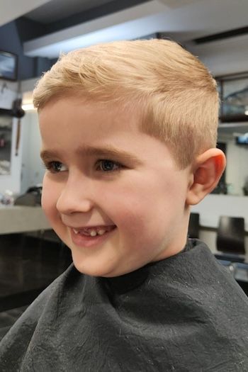 Young blonde boy with a crew cut, short sides with a subtle taper and slightly textured top, smiling in a barber chair.