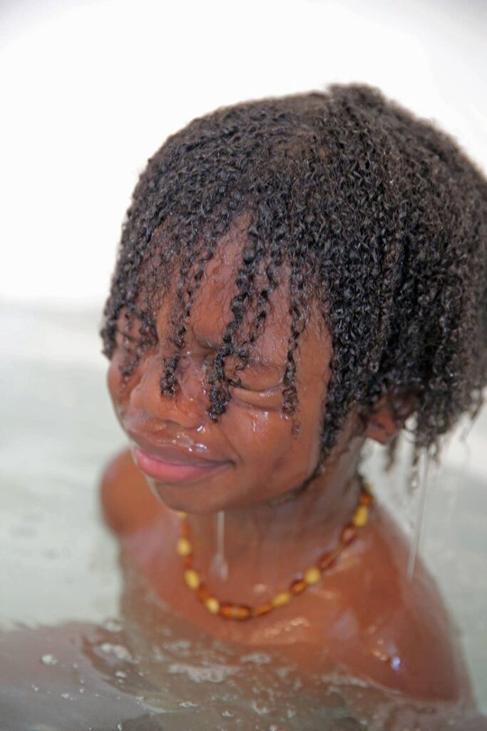 A child rinses under running water with hair sectioned into wet twists, showing the final rinse step of a wash day routine.