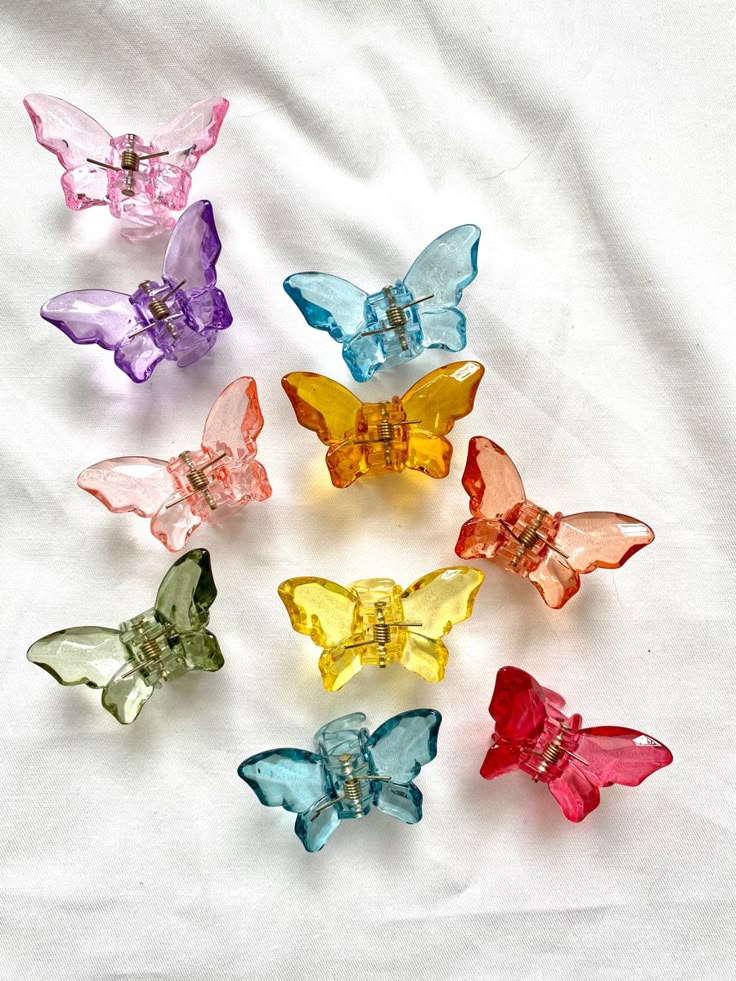 An assortment of translucent acrylic butterfly hair clips in pink, purple, blue, orange, green, and yellow, arranged on a white fabric background.