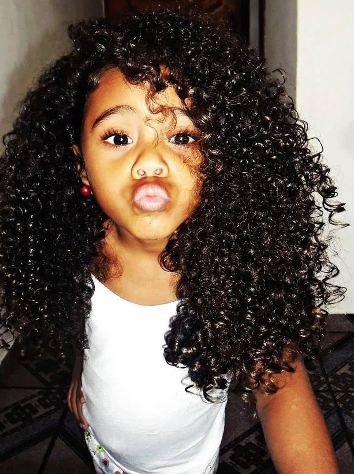 Girl with long, voluminous curly hair making a playful pucker face, wearing a white tank top.