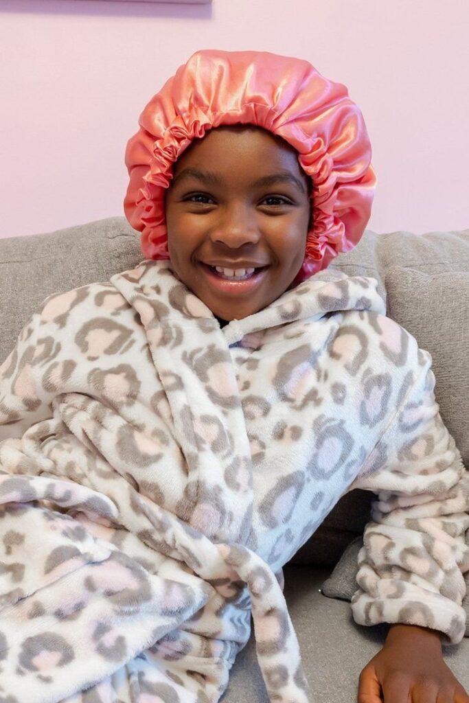 Smiling girl wearing a pink satin bonnet and a leopard print robe, sitting on a couch.