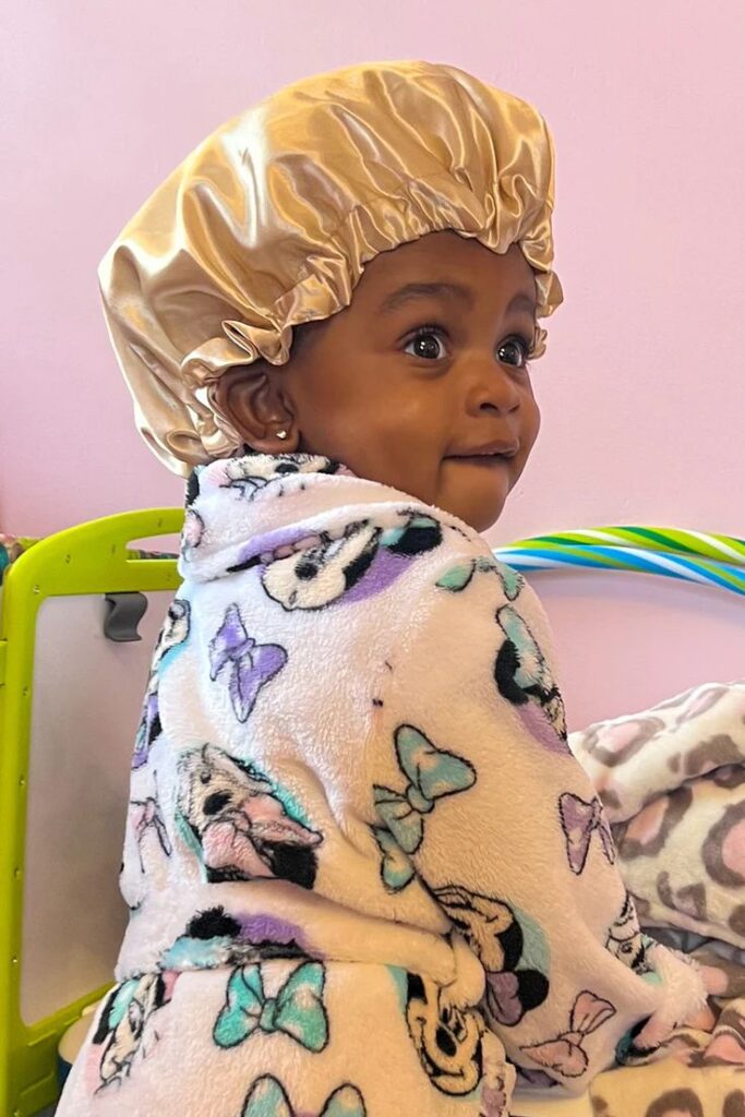 A baby wears a satin bonnet to bed, dressed in patterned pajamas, showing how a bonnet protects curls overnight.