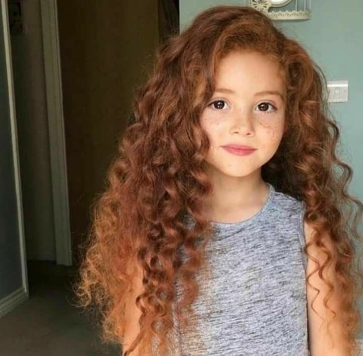 Young girl with long, loose 3A curls in a warm auburn shade, soft freckles, and a gray tank top, standing indoors near a doorway.