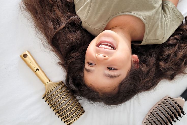 A child lying down and laughing, surrounded by long wavy hair with a gold detangling brush placed beside her.