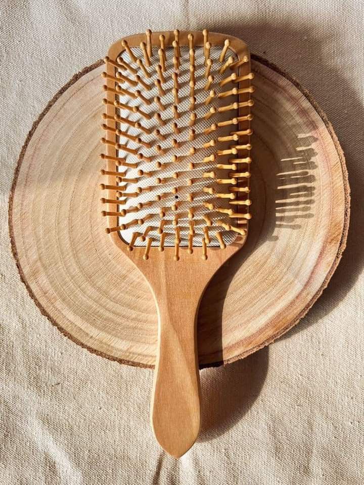 A wooden paddle brush with widely spaced pins, shown on a neutral wooden background.