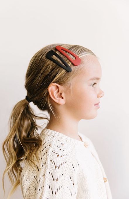 A young girl with wavy blonde hair in a low ponytail, wearing red and black snap clips near her hairline.