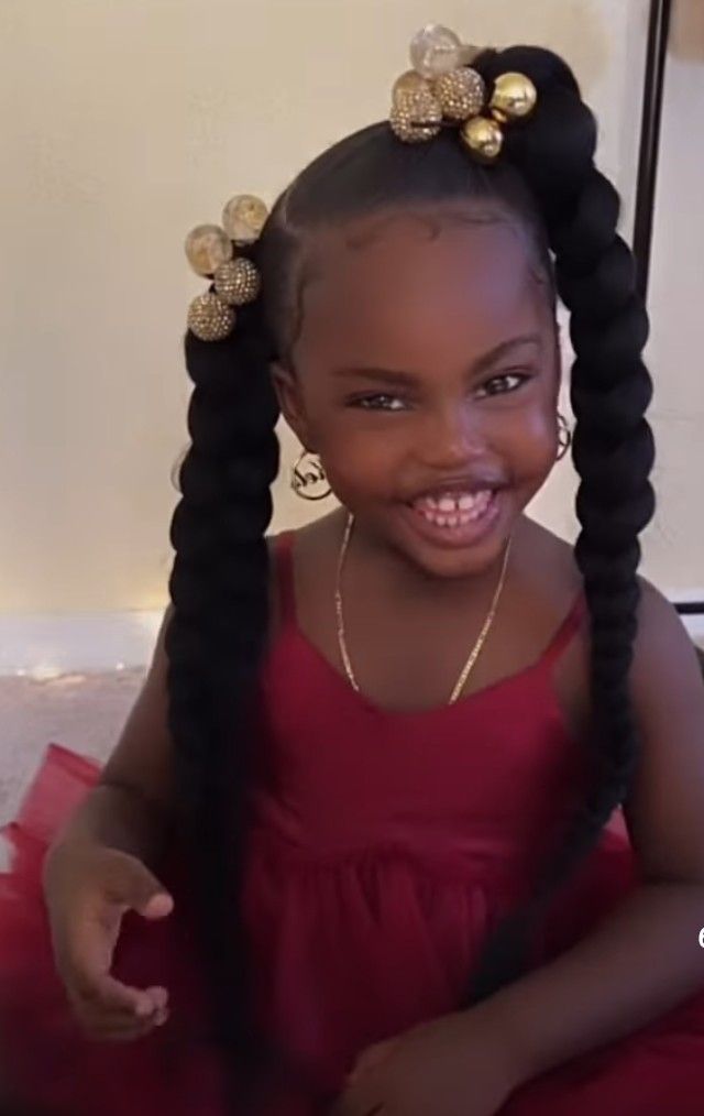 Little girl with twisted pigtails and gold bead accessories, smiling in a red dress."