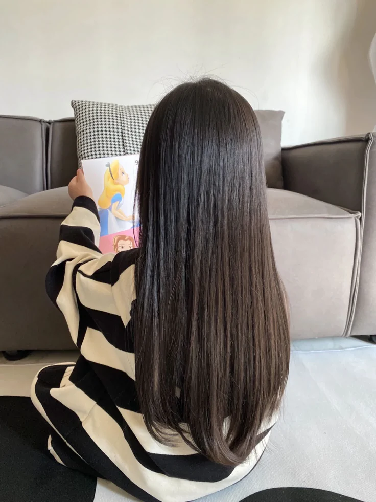 Back view of a child with long, sleek straight dark hair reaching past the waist, sitting cross-legged while reading a picture book