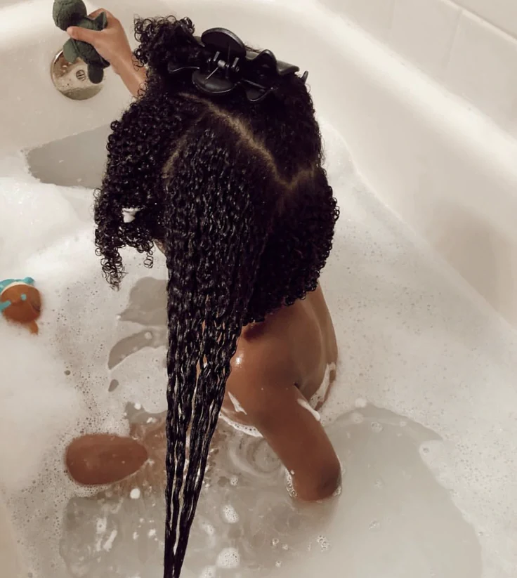 Back view of a child sitting in a bubble bath with long, defined curly hair parted down the middle, hair clip holding the top section back.