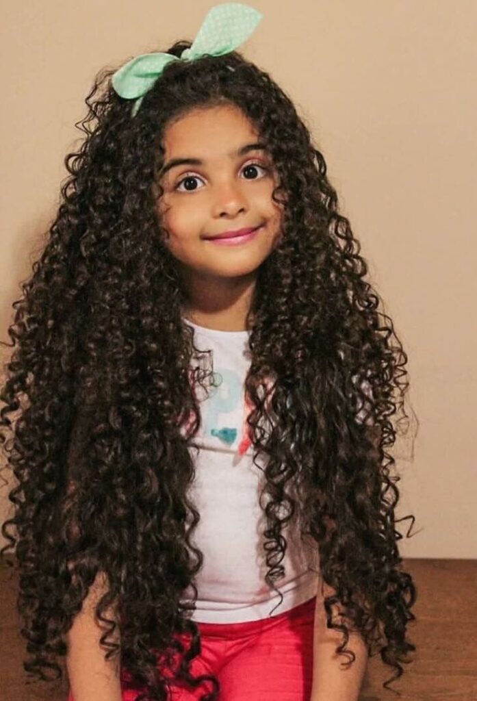 Young girl with long, defined curly hair styled with a green bow, smiling while wearing a white top.