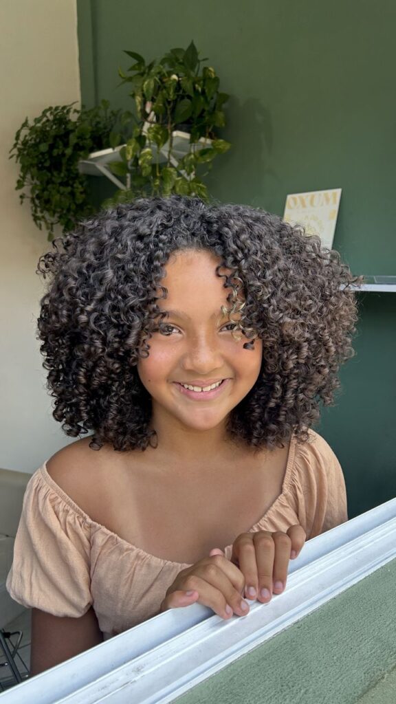 Young girl with tightly coiled 3C curls smiling near a window, wearing an off-shoulder tan top.