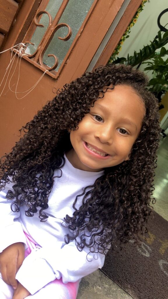  Smiling girl with long, defined curly hair sitting near a wooden door with plants in the background.