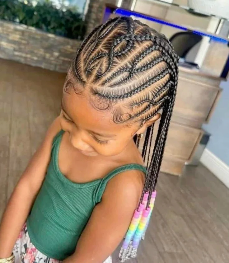 A child wears Fulani-inspired feed-in braids with a decorative crisscross pattern at the crown, finished with rainbow beads.
