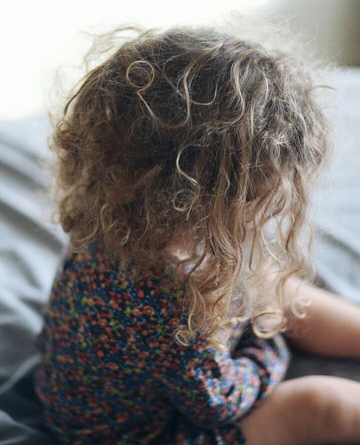 Back view of a child's dry, frizzy curly hair while sitting on a bed."