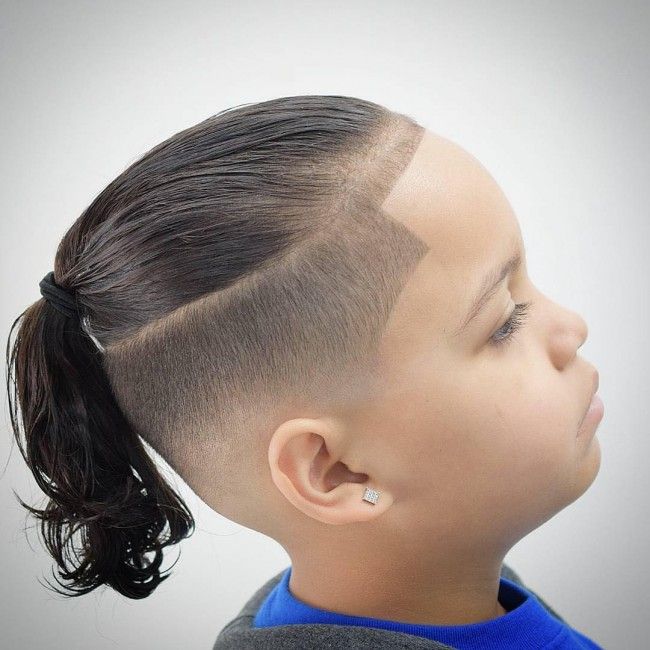 Side profile close-up of a young boy with an undercut haircut, hair on top styled back into a small ponytail with a sharp part line, blending into closely shaved sides.