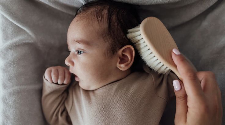 A parent brushing a newborn baby's hair with a soft-bristle brush while the baby lies on a soft blanket."