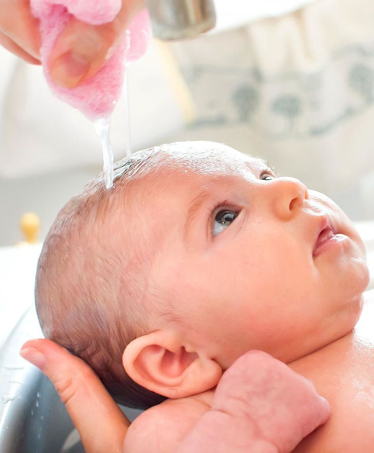 A parent rinsing water over a baby's soapy scalp during bath time, with the baby looking up calmly."