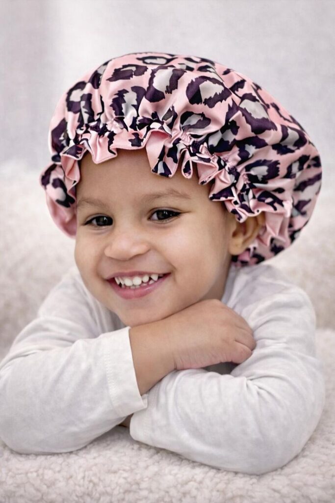 A smiling child wearing a pink and leopard print satin bonnet to protect her hairstyle