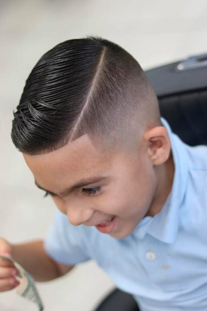 Overhead view of a young boy smiling, hair styled in a comb over with longer top hair swept to one side and a sharp part line, blending into a skin fade on the sides.