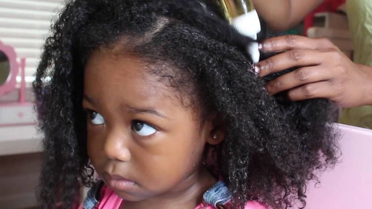 A parent applying oil to a child's scalp using a spray or applicator bottle, parting through thick curly hair."