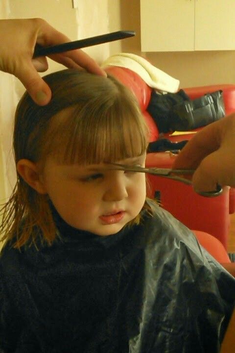 A toddler getting their bangs trimmed with scissors during a salon haircut, wearing a black cutting cape."