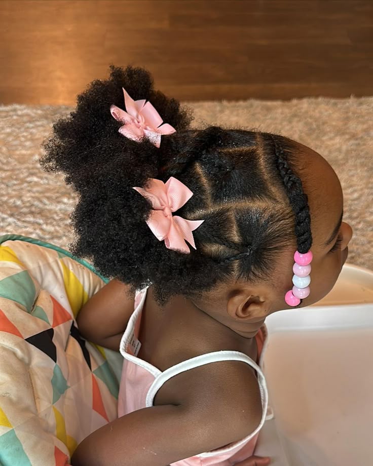 "A toddler with triangle-parted cornrows leading into a curly puff, decorated with pink bows and beads."
