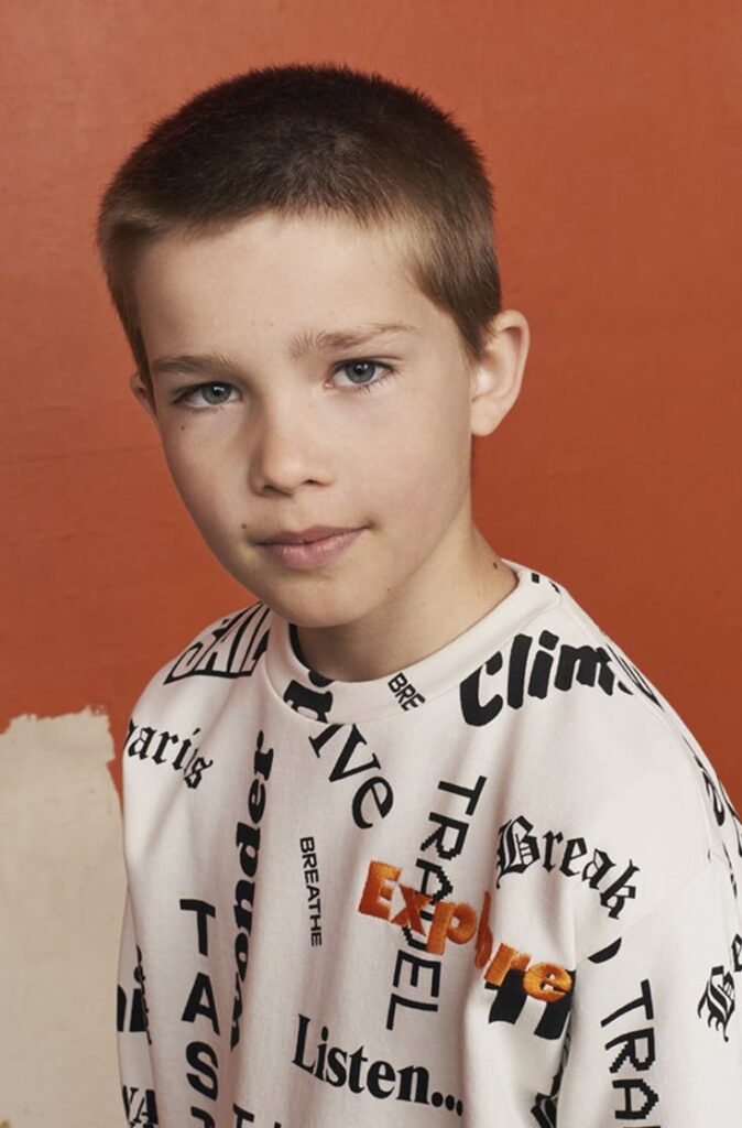 Close-up portrait of a young boy with a short, uniform butch cut haircut, wearing a graphic text-printed sweatshirt against an orange studio background.