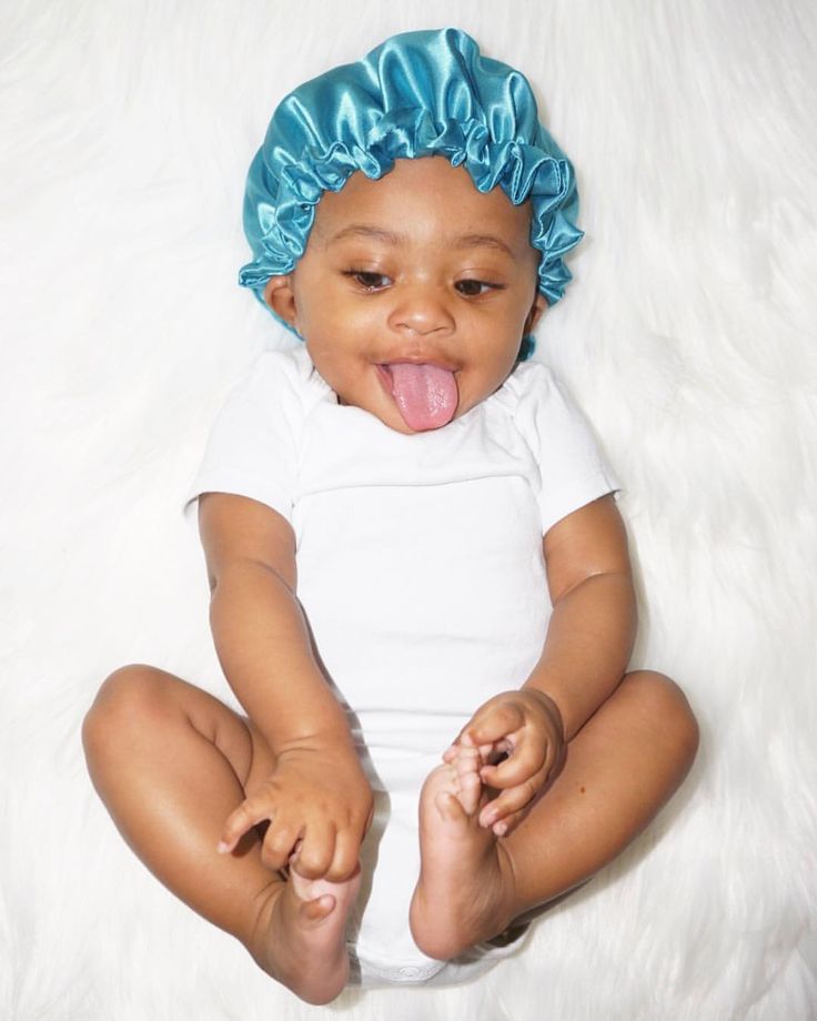 A baby lying on a fuzzy blanket wearing a teal satin bonnet, smiling with their tongue out."