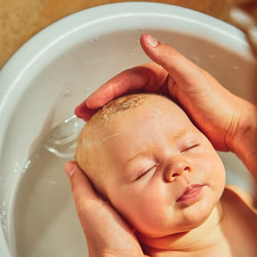 A parent rinsing water over a baby's soapy scalp during bath time