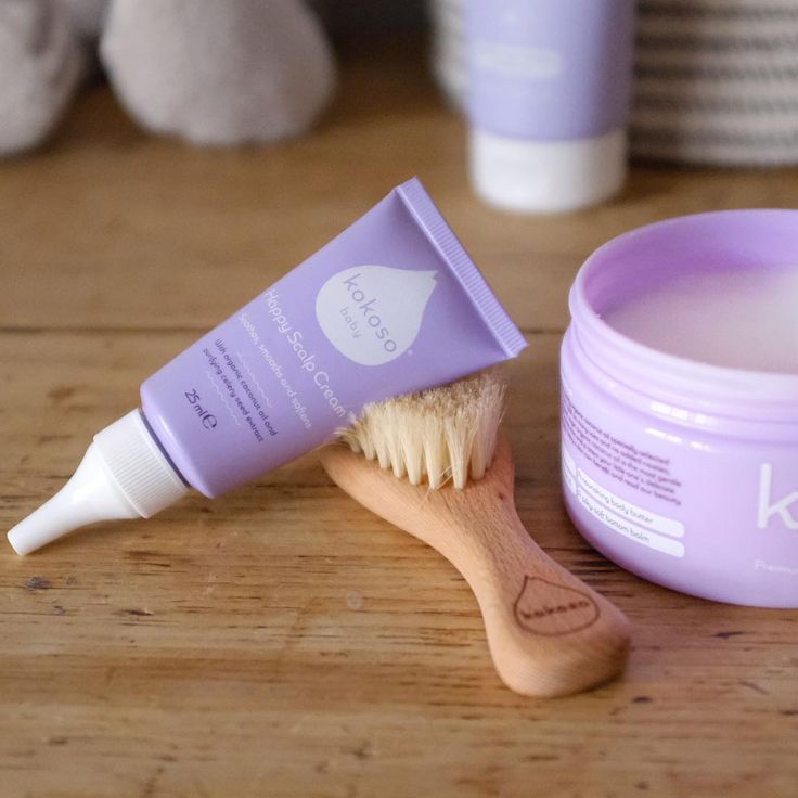 A tube of scalp cream and a soft-bristle scalp brush next to a tub of body butter on a wooden surface."