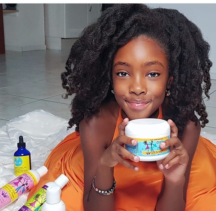 A smiling child with long, healthy curly hair holding a jar of curl cream, surrounded by other hair care products."