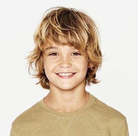 Close-up portrait of a young boy smiling, hair styled with loose, tousled surfer hair and sun-kissed highlights throughout.