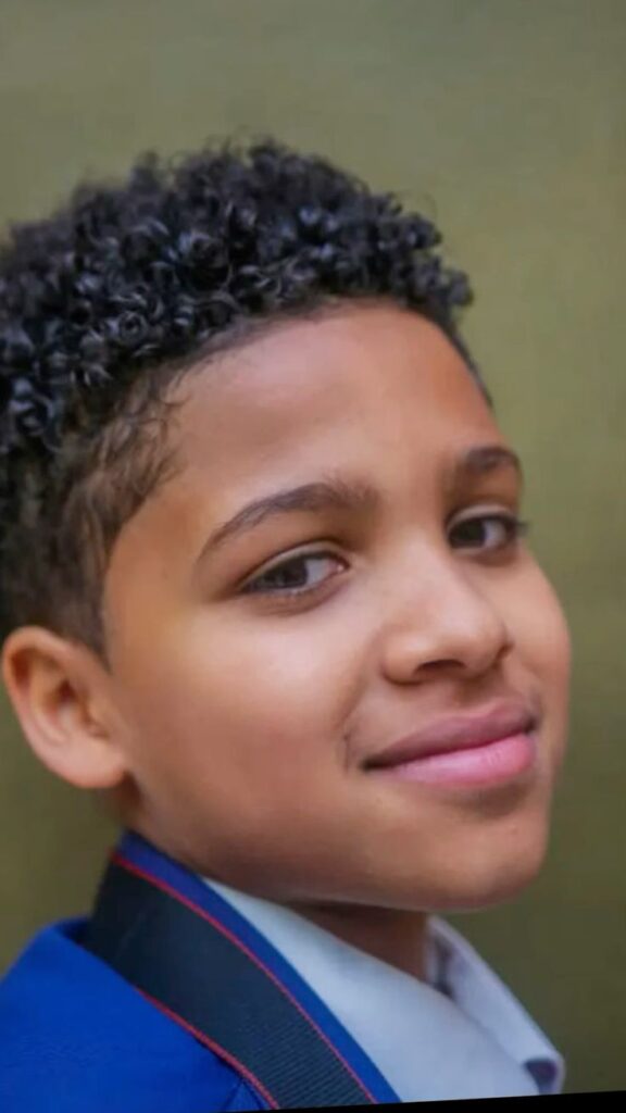 Close-up of a young boy with medium-length curly hair styled naturally, showing defined coil texture and volume on top.