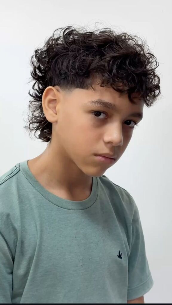 Side profile of a boy with short faded sides and longer curly length at the back, styled as a curly mullet.