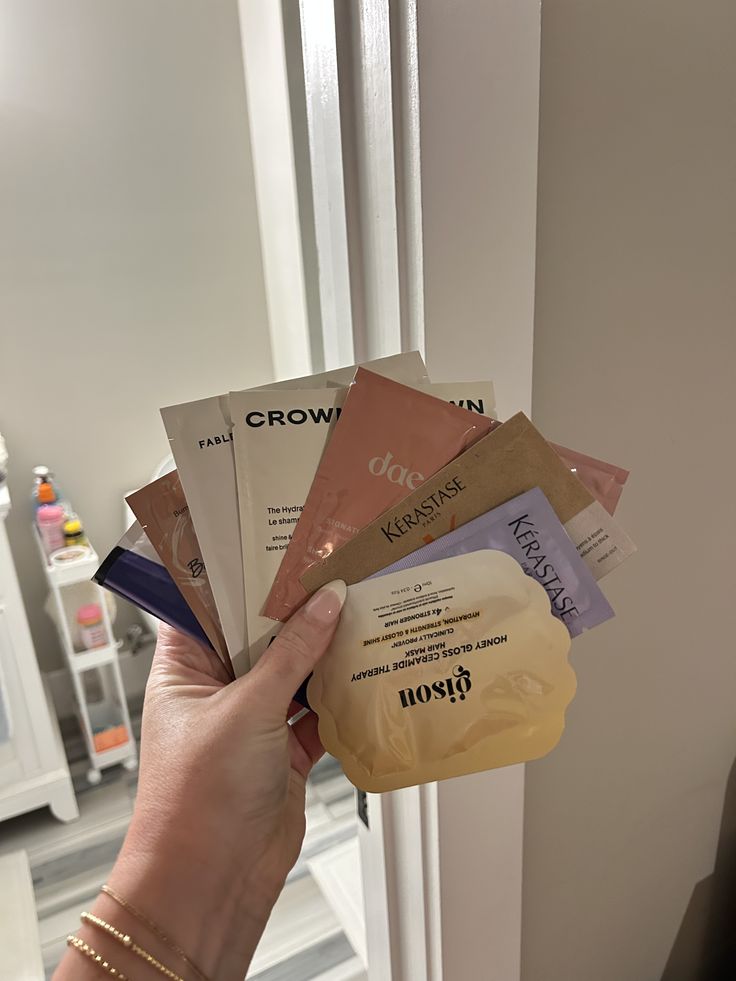 Hand holding a fan of small sample-size hair product sachets from various brands."
