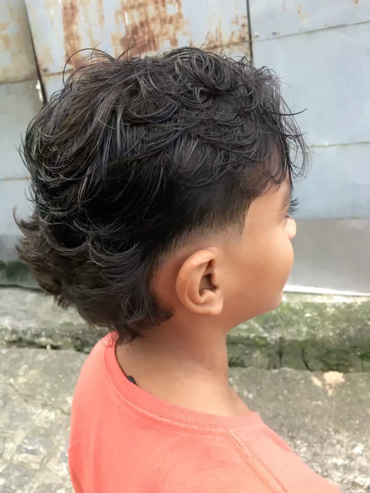 Side profile close-up of a young boy with a mullet haircut, curly hair kept longer at the back with short, faded sides.