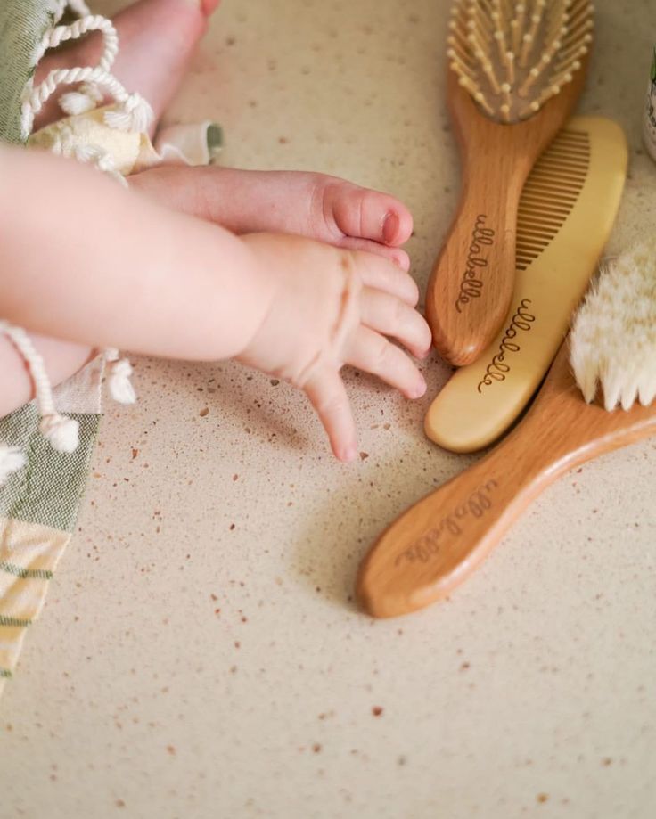 A baby's hand and foot reaching toward a set of wooden hair brushes and a comb laid on a table."