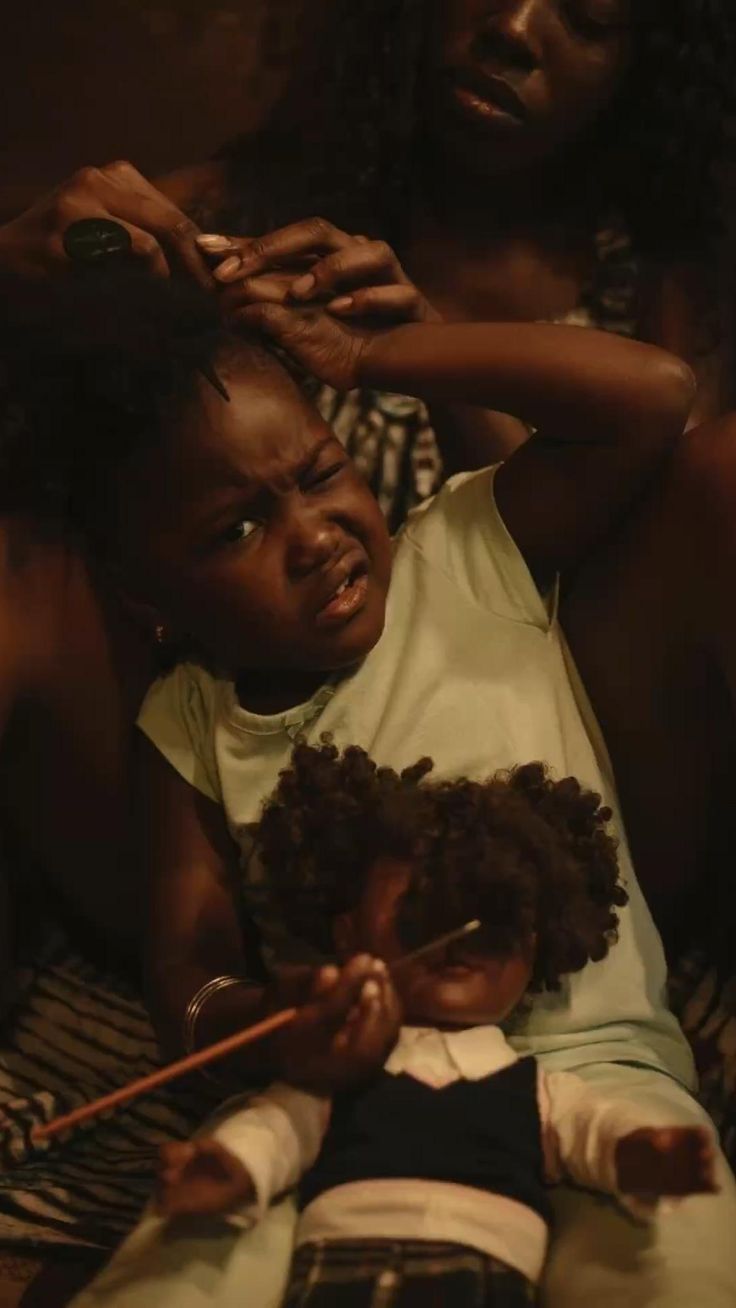 Child wincing while an adult combs her hair, holding a doll with matching curly hair.