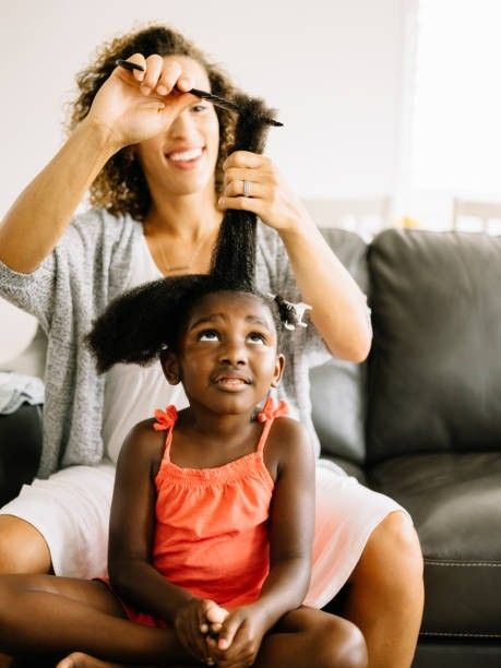 Mother detangling her smiling daughter's hair while sitting together on a couch