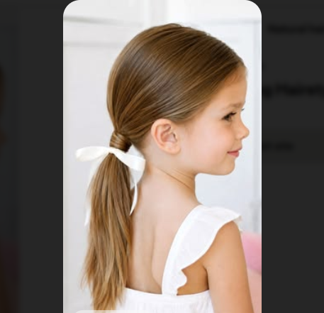 Young girl with a low side ponytail tied with a white ribbon, smiling over her shoulder